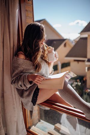 Young woman sitting with tasty drink and book on window