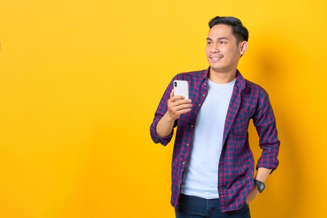 Relaxed Asian male in plaid holding smartphone mobile on yellow background