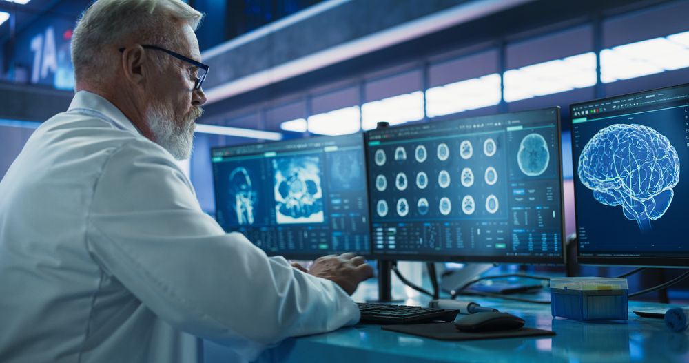 Male Neuroscientist Using Desktop Computer To Analyze MRI Scans Of Brain In Medical Research Center Laboratory.