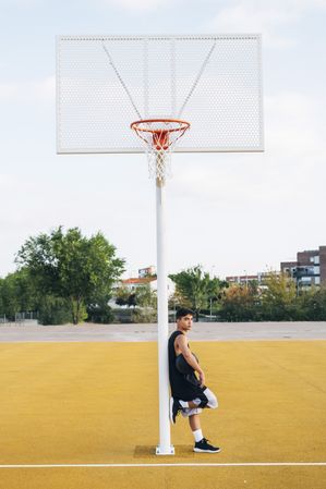 Young man leaning on basket on basketball court outdoor.