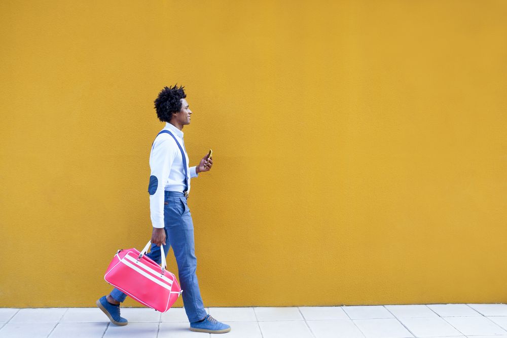 Side view of Black male with curly hair wearing shirt and suspenders walking next to yellow wall