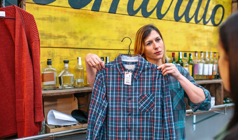 Friendly woman shop assistant with freckles showing plaid shirt to customer in store.