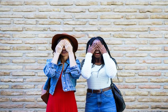 Two women smiling and covering their eyes before a brick wall