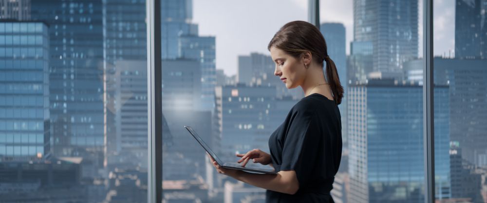 Businesswoman Standing In Her Corner Office Next To Big Window With City View And Using Laptop Computer.