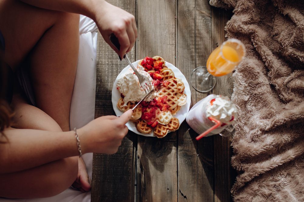 Young woman eating appetizing waffles and tasty drinks on tray on bed