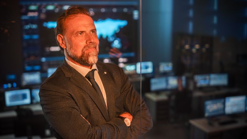 Mature Businessman in Control Room, Arms Crossed, Observing Digital Displays.