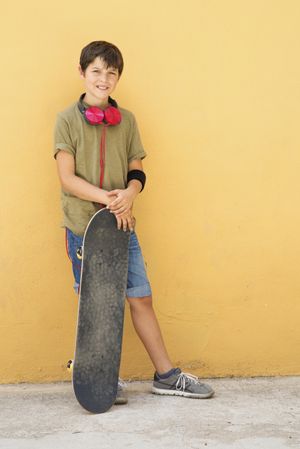 Young boy leaning on a yellow wall with headphones on neck, holding a skateboard