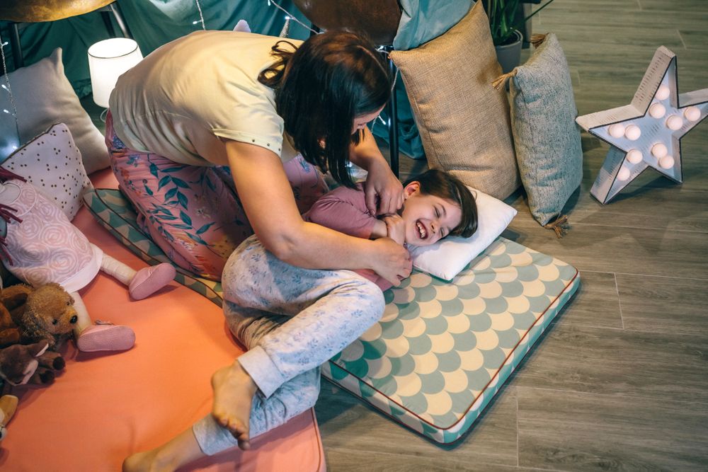 Happy mother tickling to young daughter while laughing lying over mats in night pajama party