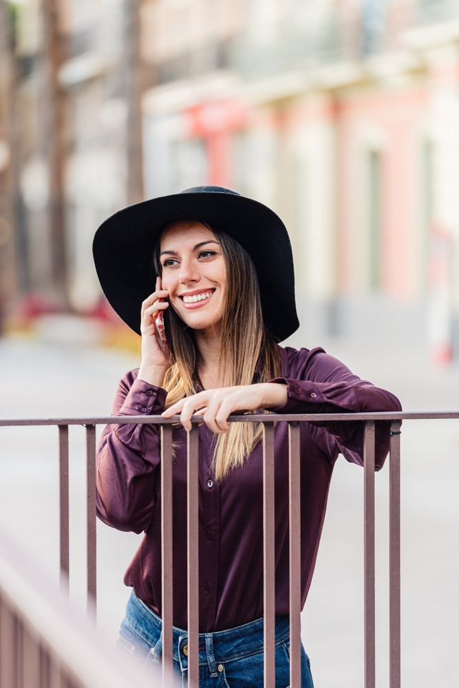 Charming woman in trendy outfit talking on smartphone and smiling at city street
