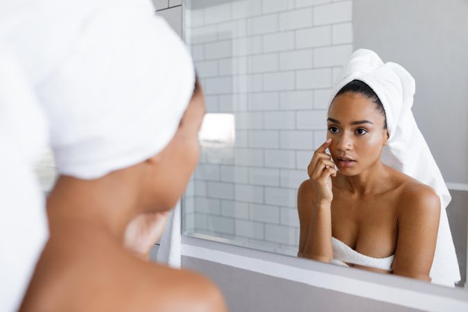 Serious Black woman in towel touching her face in the mirror