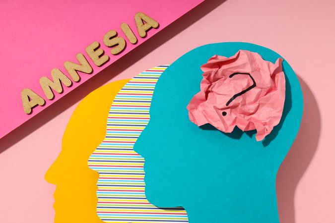 Flat lay of paper cut outs of colorful head on pink background with the word “Amnesia”