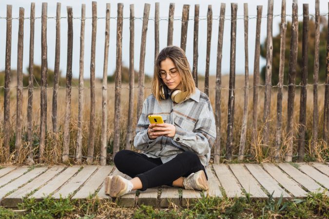 Young female millennial resting on stone border while using mobile phone at roadside
