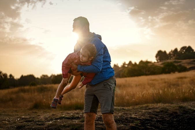 A father playfully lifting his daughter in the air like an airplane at sunset.