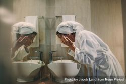 Side view of woman in bathrobe washing her face in bathroom sink and looking into the mirror 4ZP630