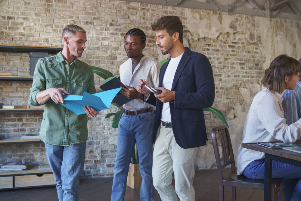 Three male colleagues discussing with folder in hand while walking, with other people sitting at desks in the background