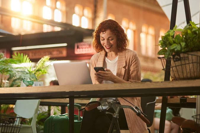 Cheerful young woman working on laptop in cafe and browsing phone