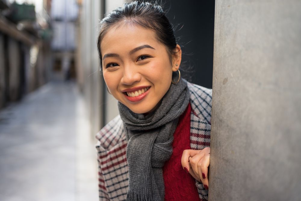 Joyful Asian resting female in casual clothing looking out modern wall of building at city street