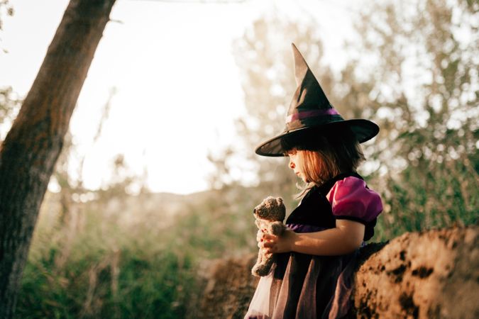 Quiet girl in witch hat with teddy bear in the forest