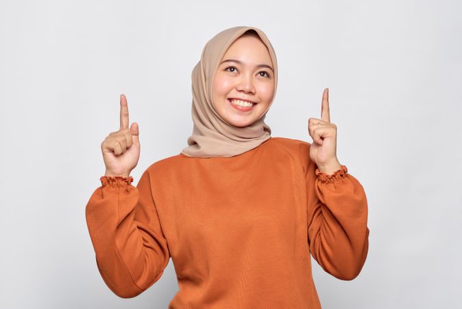 Smiling Muslim woman in headscarf and orange sweater pointing upwards with good idea