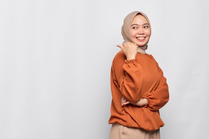 Smiling Muslim woman in headscarf and orange sweater pointing behind her with thumb and winking