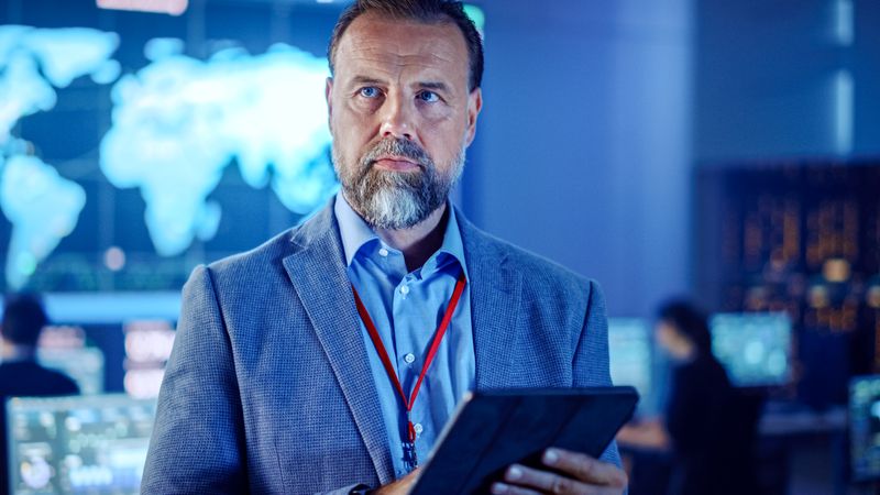 Bearded Man in a Blue Suit Analyzes Data on a Tablet in a Futuristic Control Room