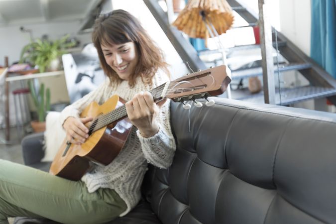 Smiling female on sofa enjoying playing music of her acoustic guitar in living room