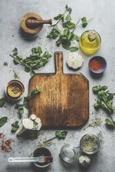 Empty wooden cutting board on grey concrete kitchen table with herbs ,spices, garlic,oil and mortar and pestle.