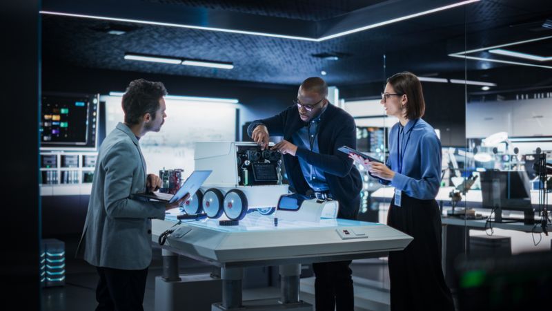 Industrial Robotics Specialists Working on a Mobile Robot, Using a Tablet Computer and Laptop. Black Male Engineer In a Lab Discussing an Automated AI Robotic Delivery Assistant with His Young Team