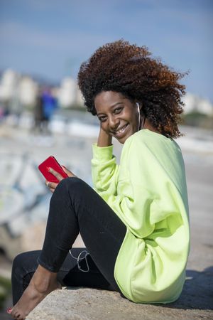 Smiling female with curly hair in bright green shirt sitting listening to music outside