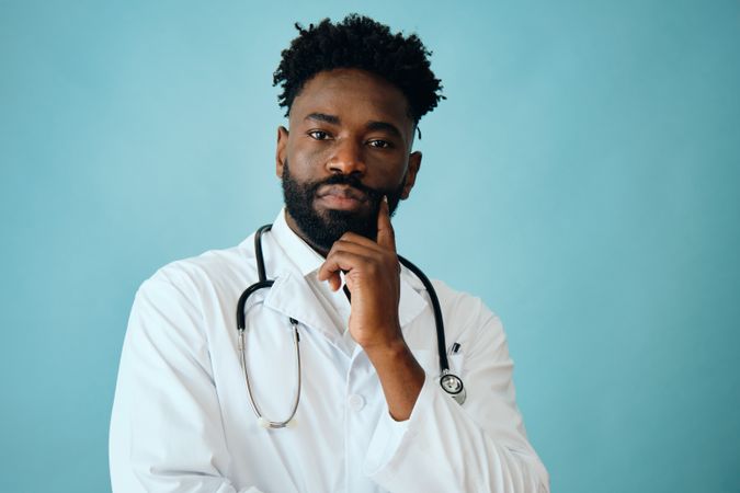 Black male doctor in blue studio with hand to chin