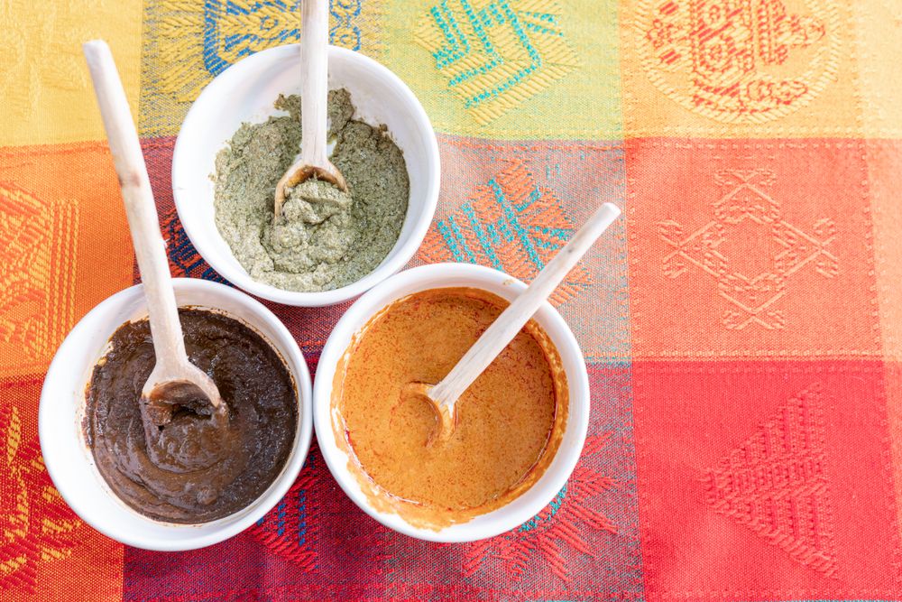 Three bowls with red, green and yellow mole sauce with a wooden spoon on top of a colorful traditional tablecloth