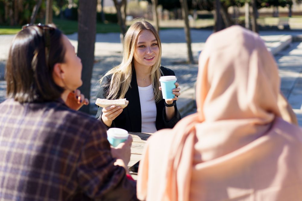 Blonde woman holding sandwich and coffee cup chatting with her friends outside