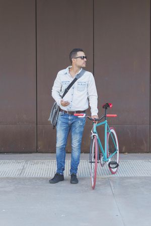 Male in sunglasses standing with red and green bicycle and looking around, vertical