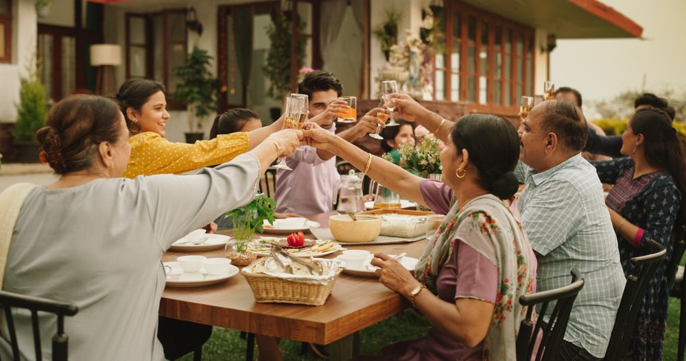 Happy Indian Family Celebrating: Group of People Gathered Together at the Table Raise Glasses To Make a Toast and Clink Glasses.