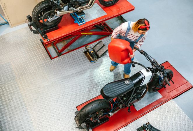 Mechanic woman with jerry can filling fuel tank of motorcycle on factory