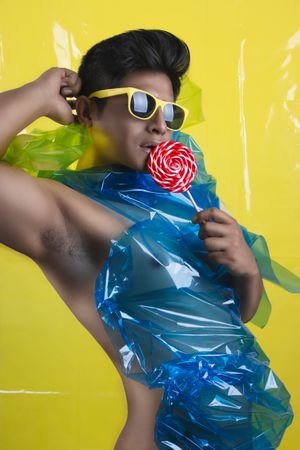 Sexy bare-chested man with blue plastic on chest wearing yellow framed sunglasses holding lollipop