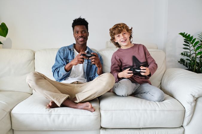 Cheerful Black man and young white boy with gamepads resting on sofa