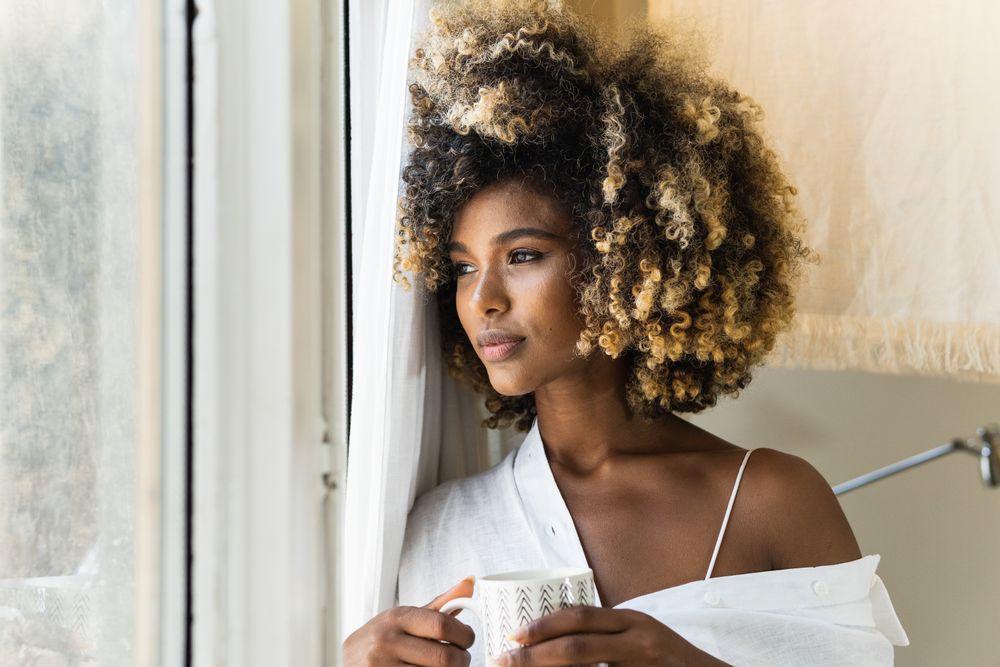 Dreamy Black woman with cup of drink near window