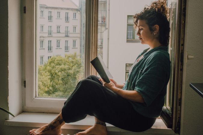 Interested woman reading book at window sill at home