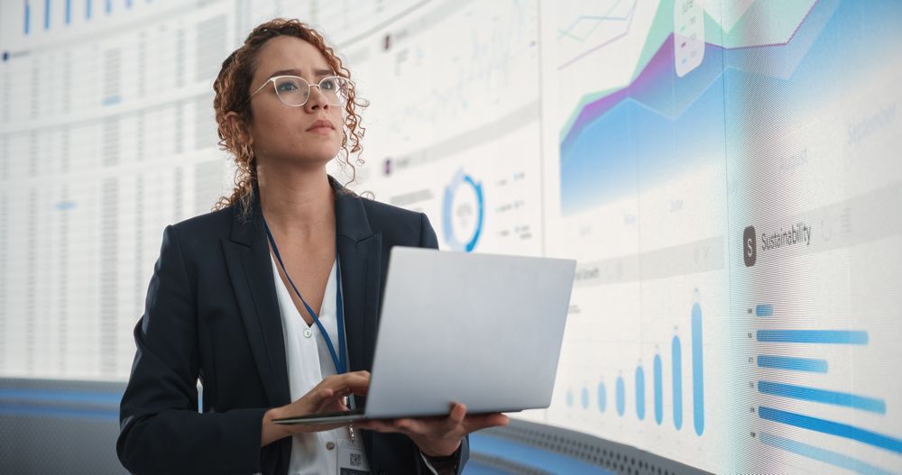 In the Monitoring Office: Successful Hispanic Businesswoman Holding Laptop Computer And Analyzing Data On Big Digital Screen With Graphs and Charts.