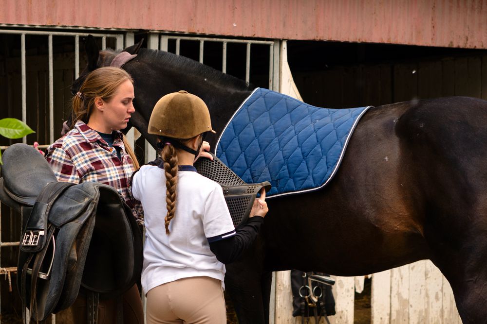 Instructor helping a girl position a saddle on a horse at a stable.
