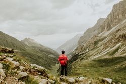 Person with backpack and hiking sticks walking on green grass field near mountain under cloudy sky