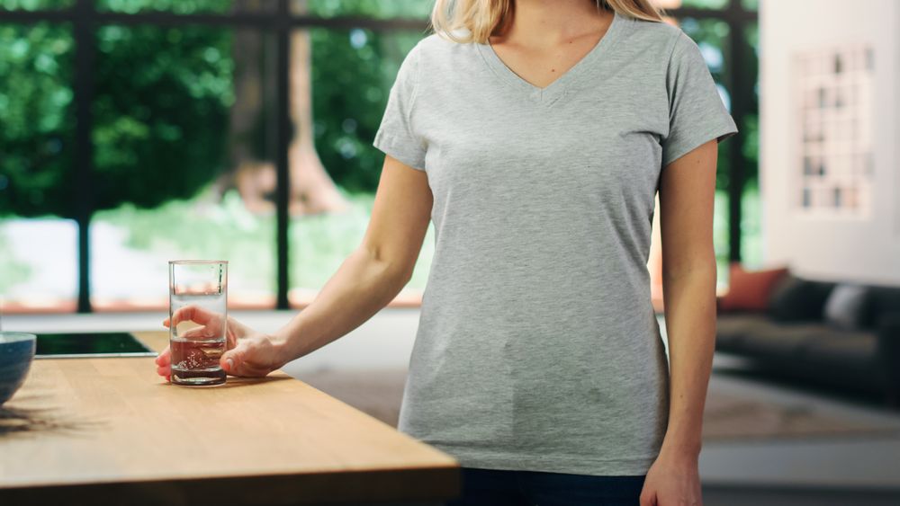 Young Adult Woman with Blond Hair Wearing Gray V-Neck T-Shirt, Holding a Glass of Water on the Table.