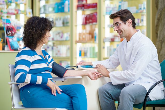Happy male pharmacist sitting and checking blood pressure of mature female customer