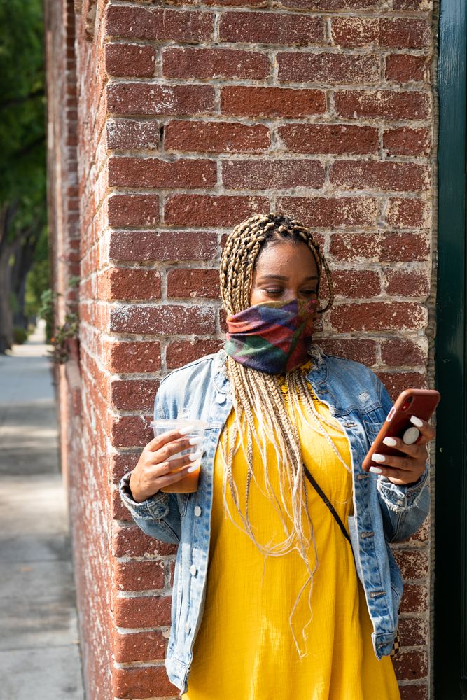 Woman with mask and long hair leaning against brick wall outside checking her mobile phone