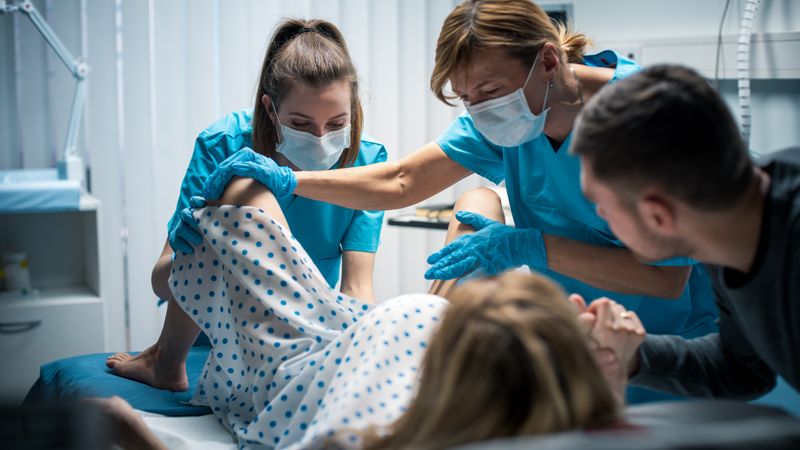 In the Hospital Woman in Labor Pushes to Give Birth, Obstetricians Assisting, Husband Holds Her Hand