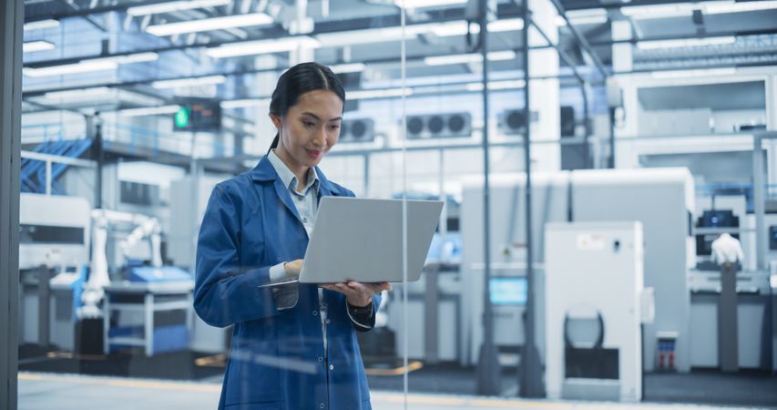 Asian Engineer Using Laptop Computer and Looking Around a Factory Facility with Equipment Producing Modern Electronic Components with Robotic Arms on Assembly Conveyor