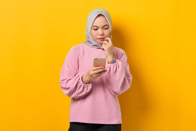 Muslim woman in headscarf contemplating something while holding smart phone with finger in her mouth