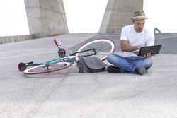 Male sitting outdoors with bicycle and using laptop