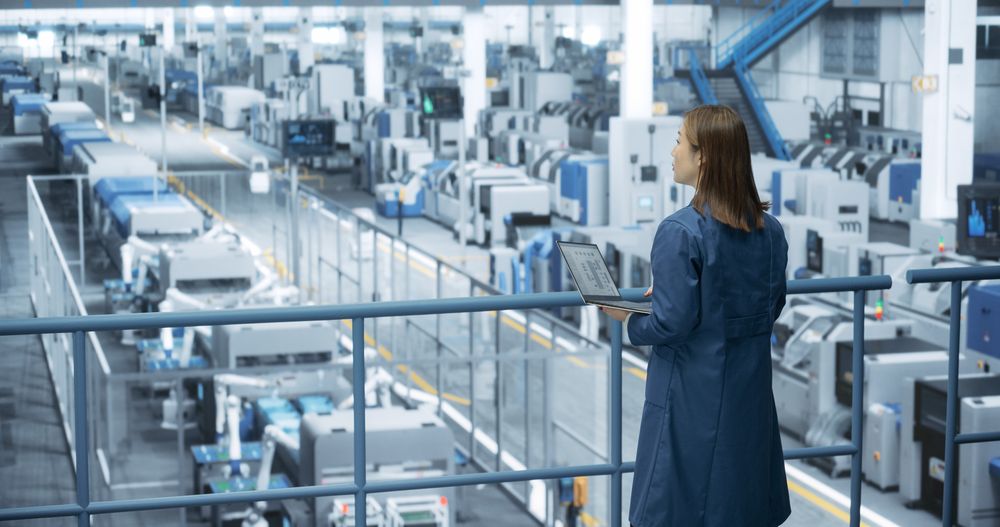 Japanese Factory Specialist Working on Laptop at a Futuristic Industrial Manufacture with Autonomous AI Robots. Project Manager Monitoring Data on a Computer, Checking Reports and Planning Research